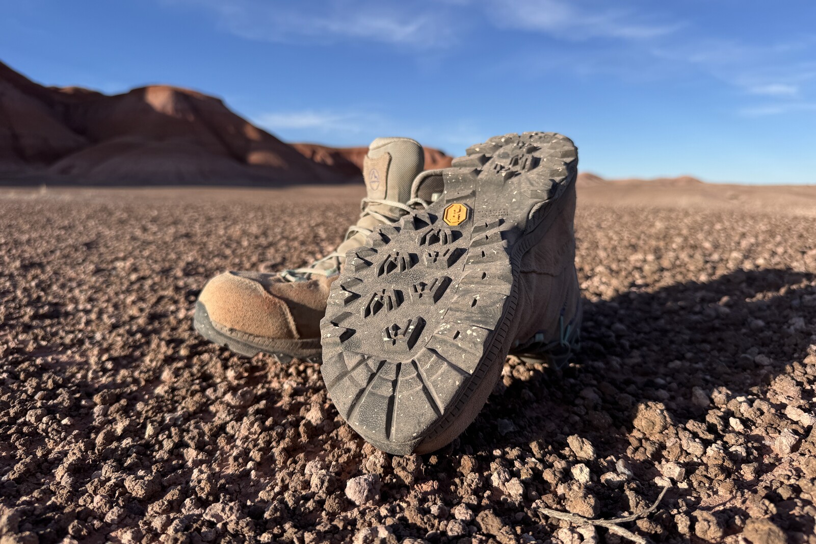 One TX Hike boot is turned over to show the tread. It is sitting on bare ground with badlands formations in the background.