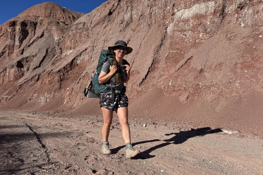 A person wearing the TX Hike boots and a large backpack walks along a wash with red badlands behind them.