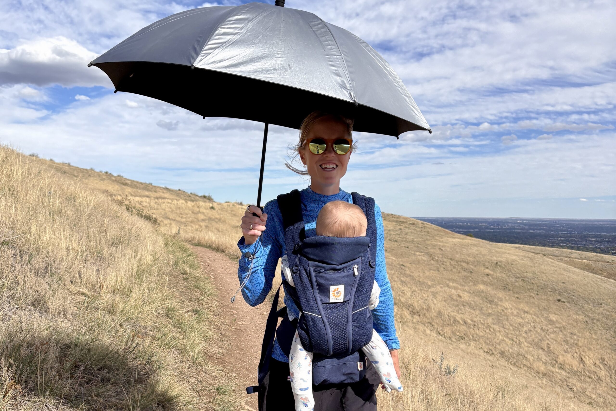 A smiling woman walks on a trail with an umbrella and a baby in a baby carrier