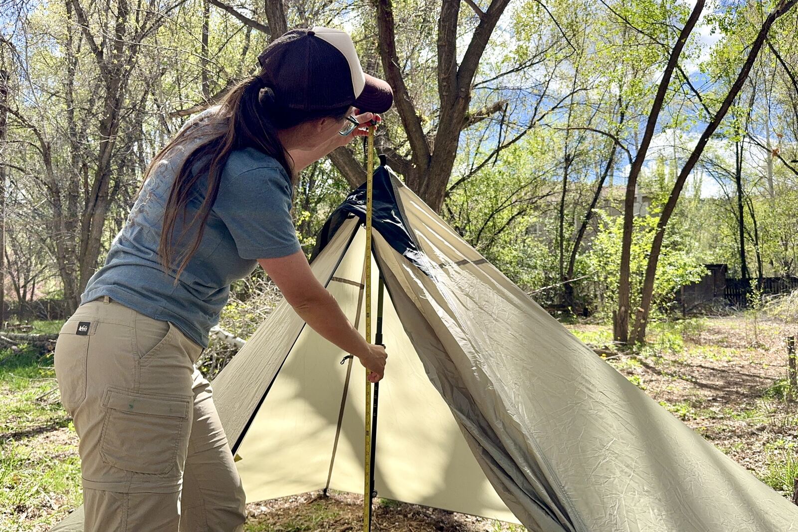 A person is holding a measuring tape up to the peak of the MLD tent. There are green trees in the background.
