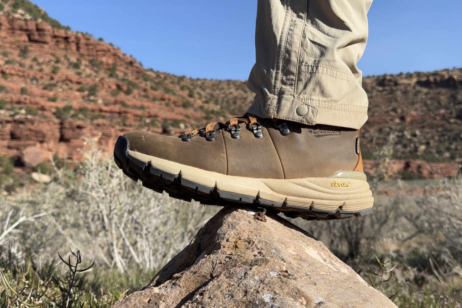 A closeup of the Danner boots as a person props their foot on a rock. There are red, rocky hills in the background.