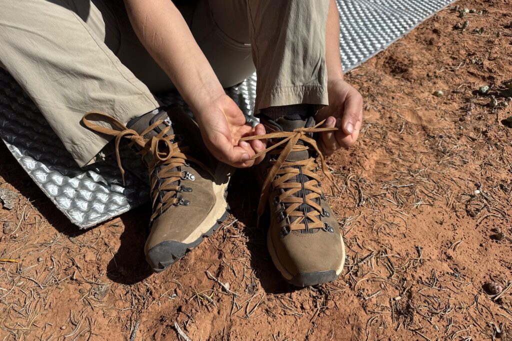 A closeup of a person tying the laces on the Danner boots. They are sitting on a foam pad on the ground.