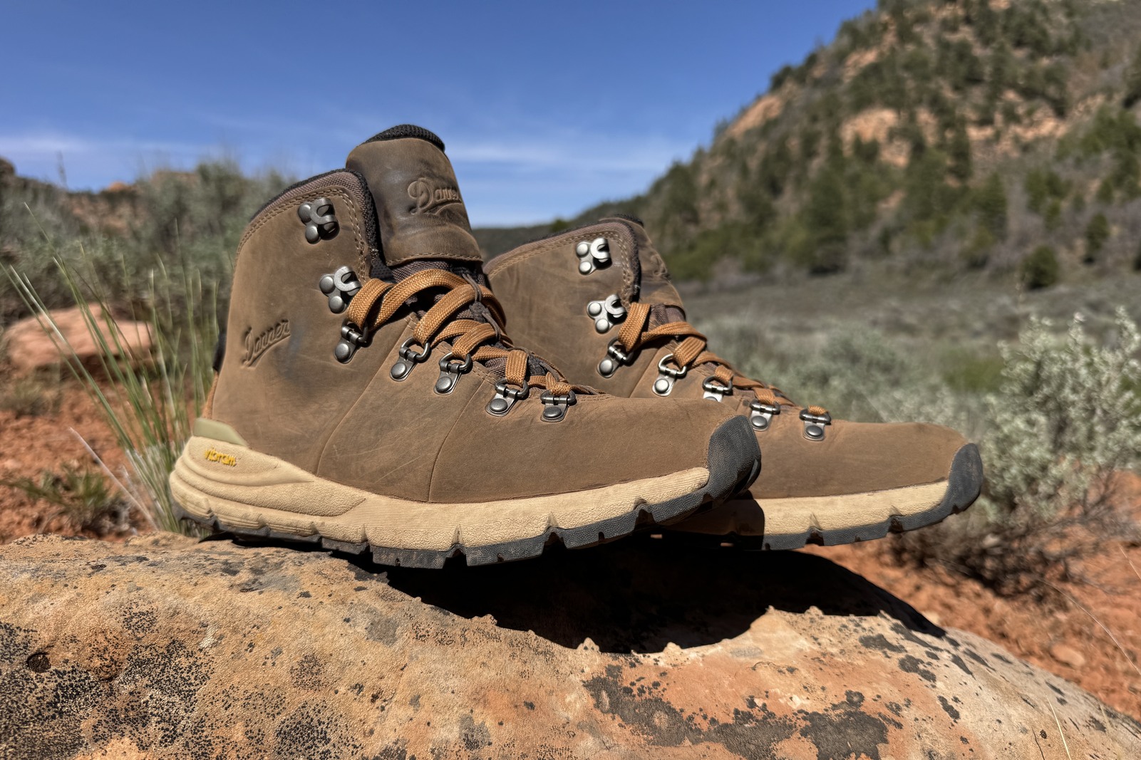 A closeup of the Danner boots sitting on a rock. There is red dirt and brushy hills in the background.