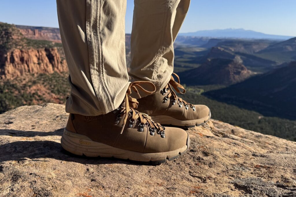 A closeup of a the Danner boots while a person is standing on a rocky overlook. There are red rock canyons and some mountains below in the background.