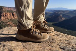 A closeup of a the Danner boots while a person is standing on a rocky overlook. There are red rock canyons and some mountains below in the background.