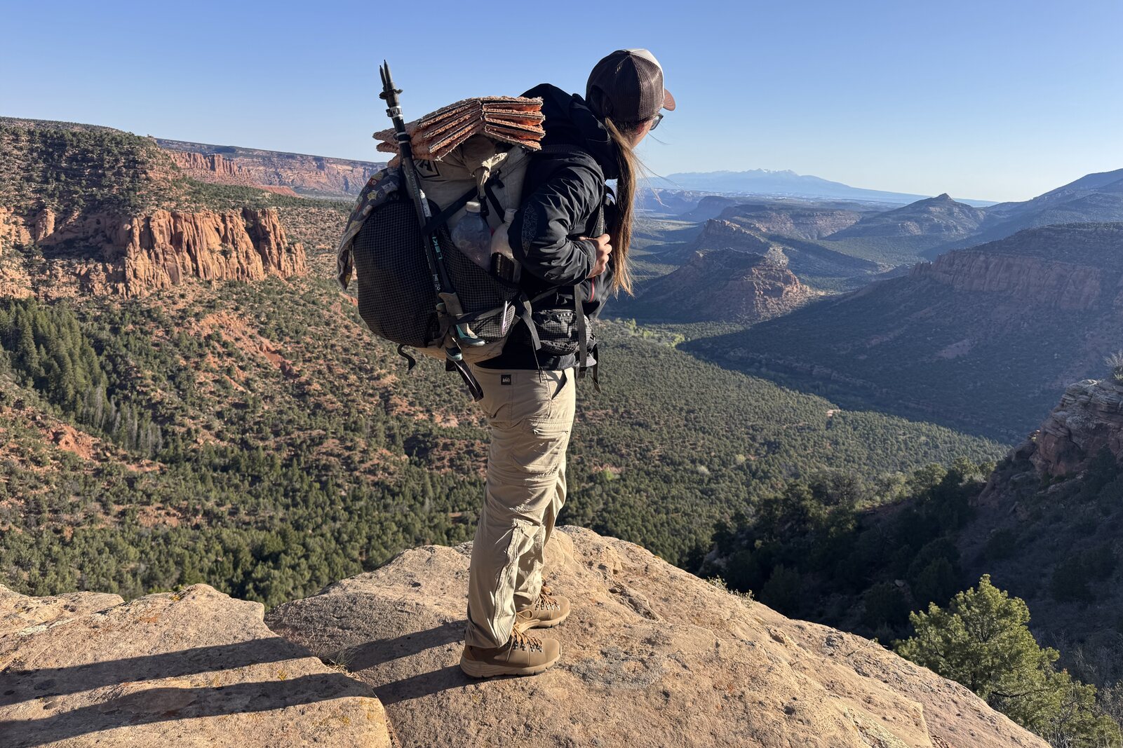 A person wearing the Danner boots and a backpack is standing on a rocky overlook admiring a view of red rock canyons and the La Sal mountains in the distance.