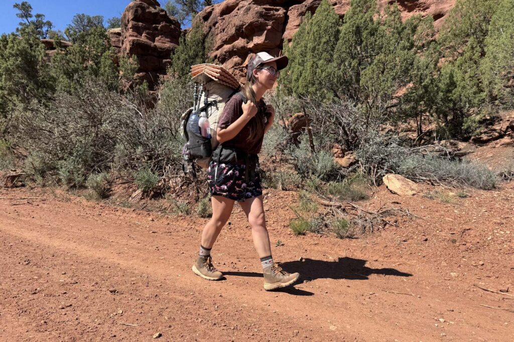 A person wearing the Danner boots and a backpack is walking down a red dirt road with red rocks and pine trees in the background.