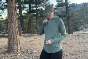 A woman wears a baseball cap and shows off the fit of a green sun hoodie in front of pine trees.
