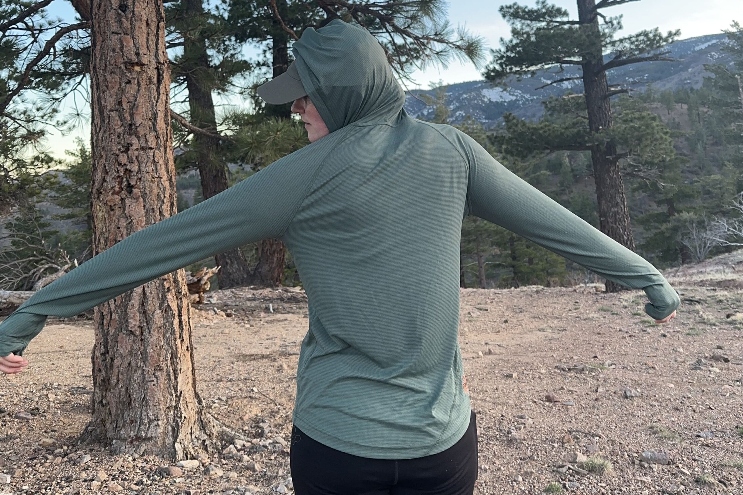 A woman stands with her back to the camera to show the fit of a shirt across the shoulders with trees in the background.