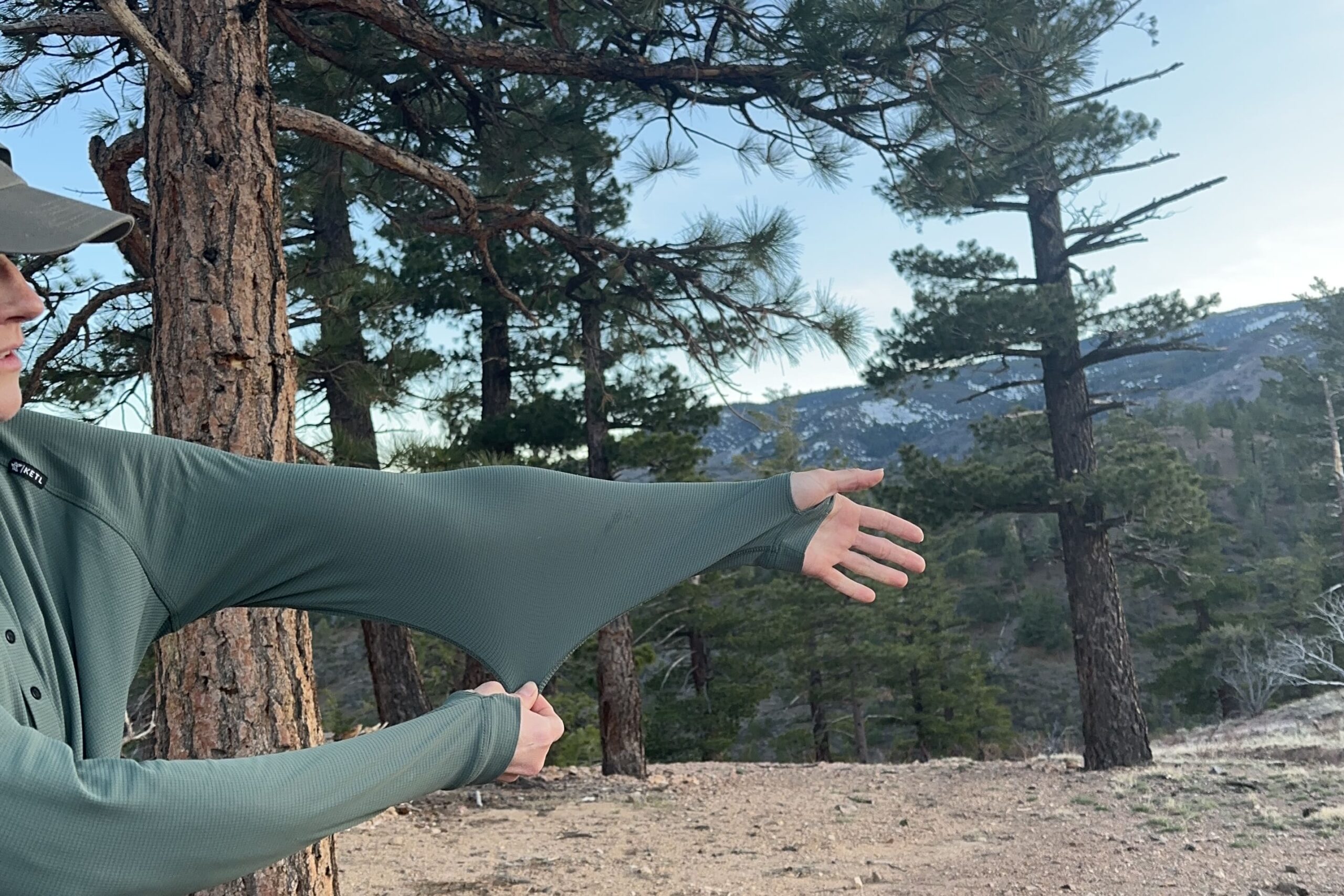 A woman holds out her arm and tugs at the fabric of her shirt to show the stretch, with trees in the background.