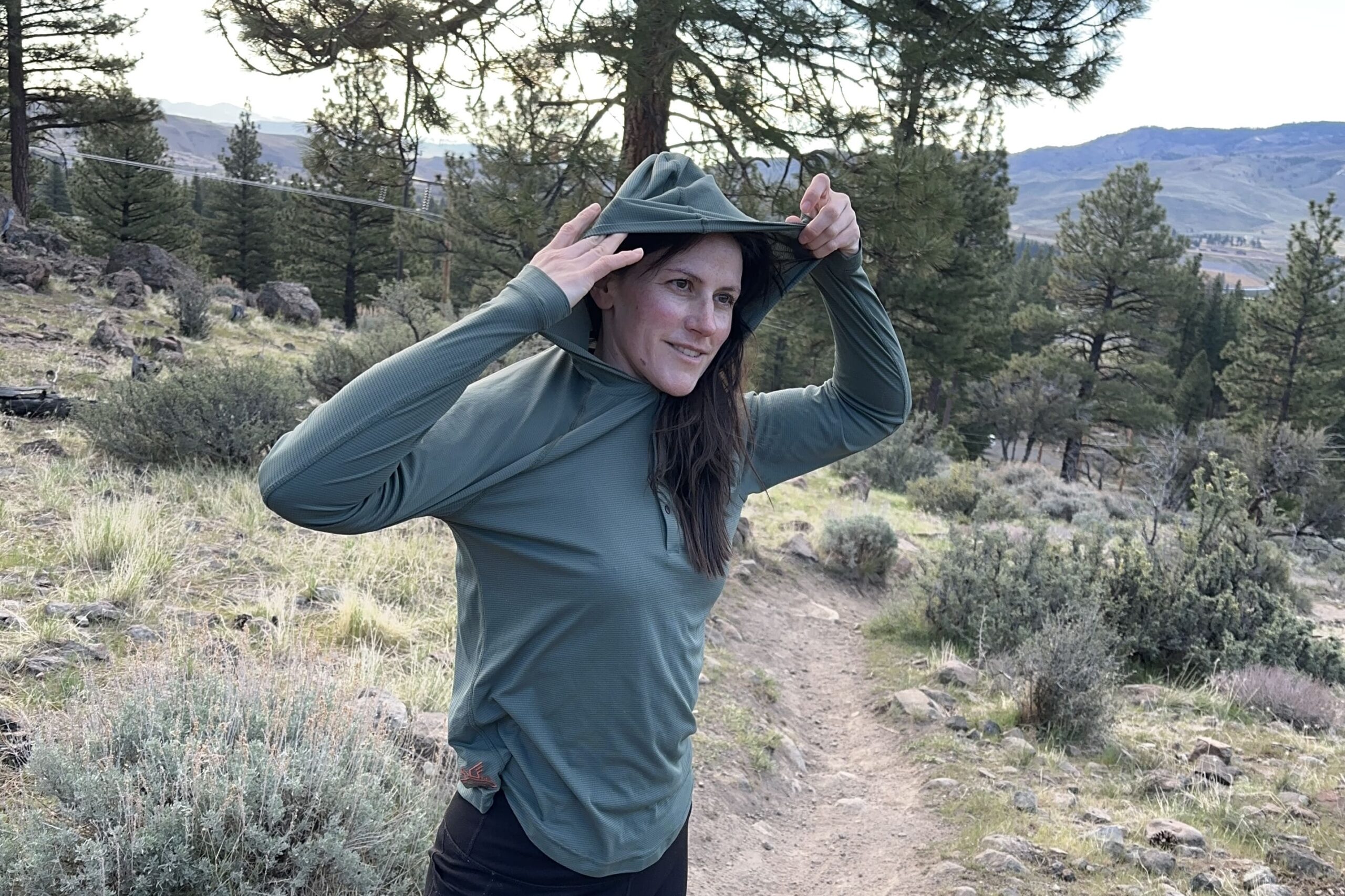 A woman adjusts the hood of her sun hoody with a high desert landscape in the background.