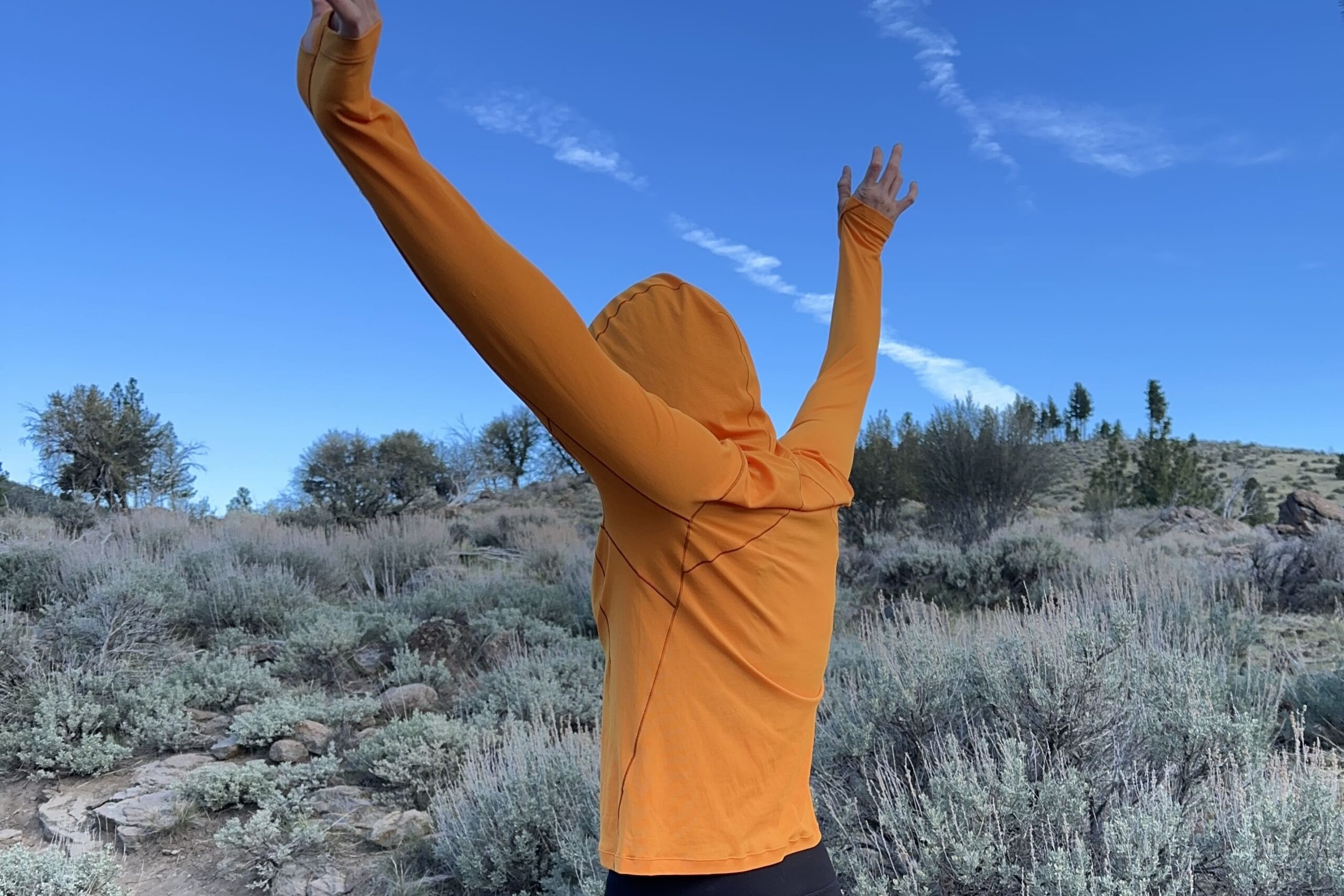 A woman in a bright orange sun shirt stands with her back to the camera with her arms raised in the air.