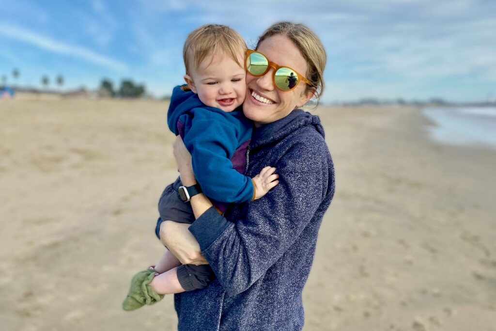 A smiling woman hugs a toddler on a beach