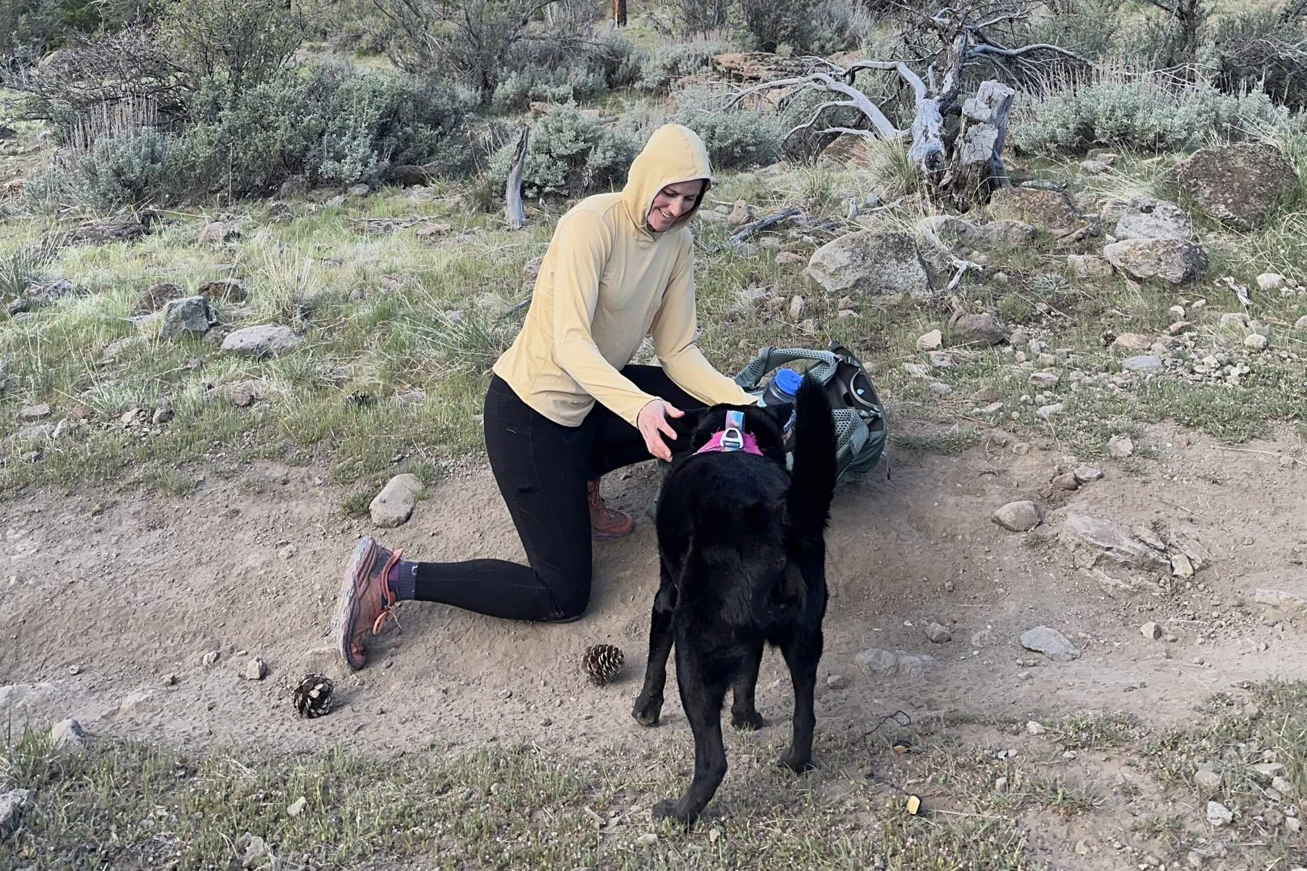 A woman in a yellow sun shirt kneels down to pet her black dog.