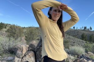 A woman in a yellow long sleeve shirts holds her arms above her head to shade her eyes as she looks into the distance.