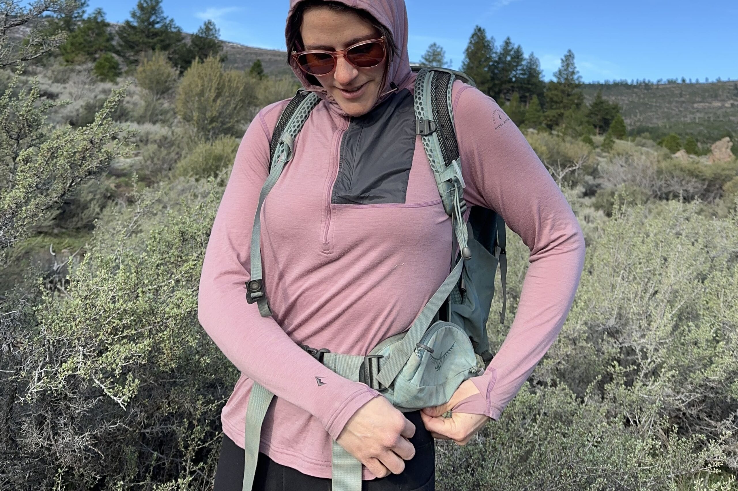 Woman wearing a pink shirt and day pack on pulling shirt corner down from under the pack belt.