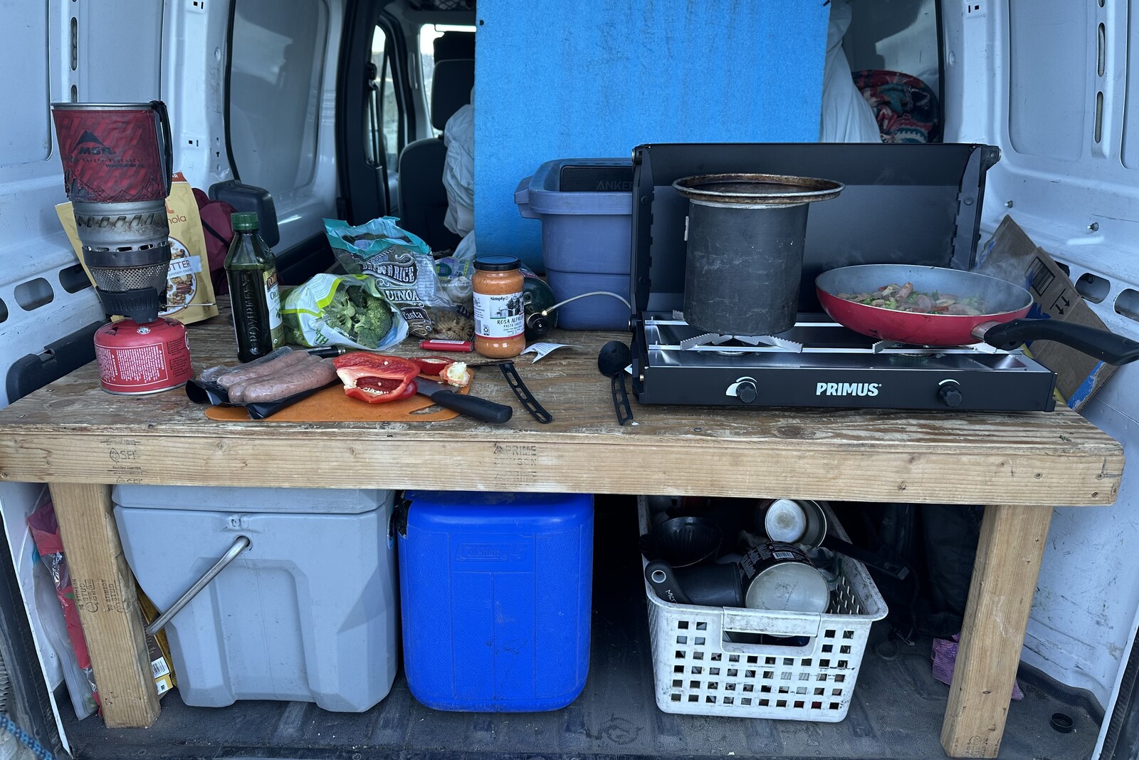Inside the back of a small camper van, a kitchen scene shows a double-burner stove with a pot and pan and a cutting board with veggies and chicken sausage.
