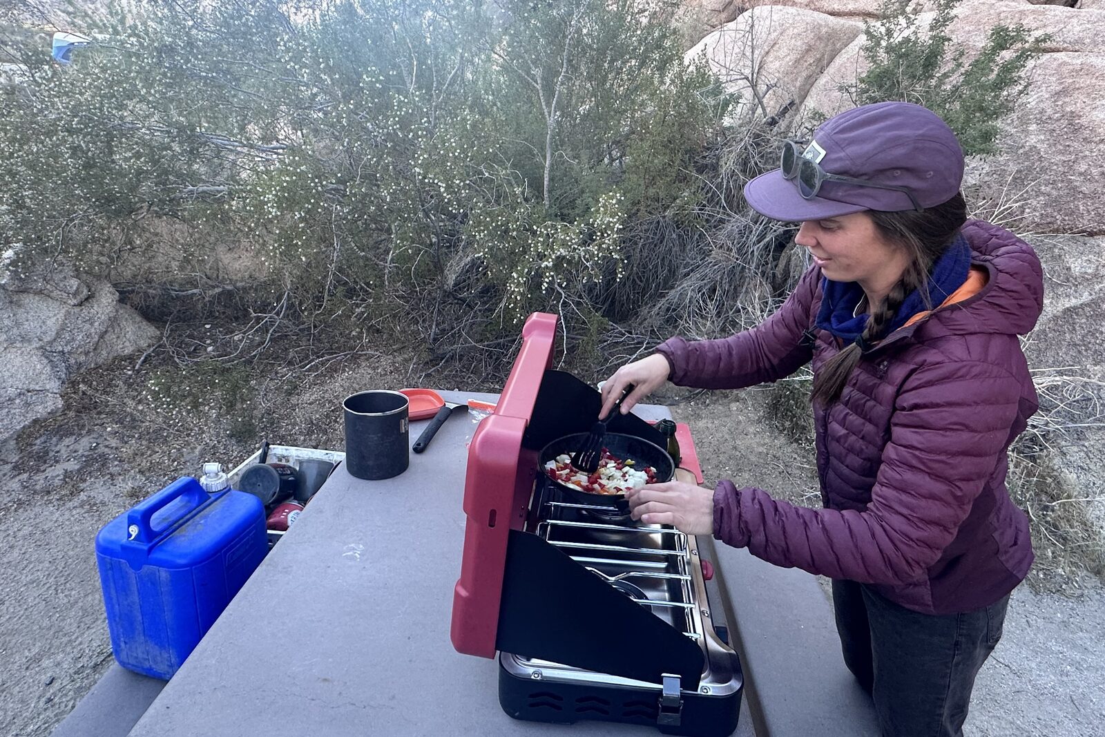 Side view of a woman cooking veggies on a double-burner camping stove with large windscreens.