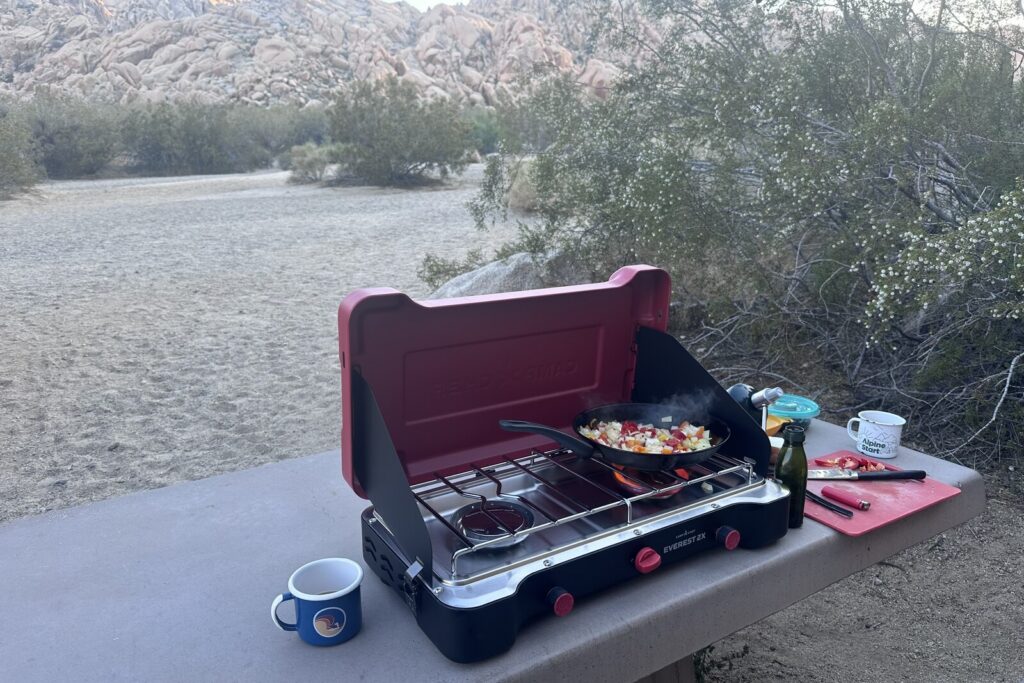 A pan of veggies cooks on a large double-burner stove sitting on a picnic table in the desert.