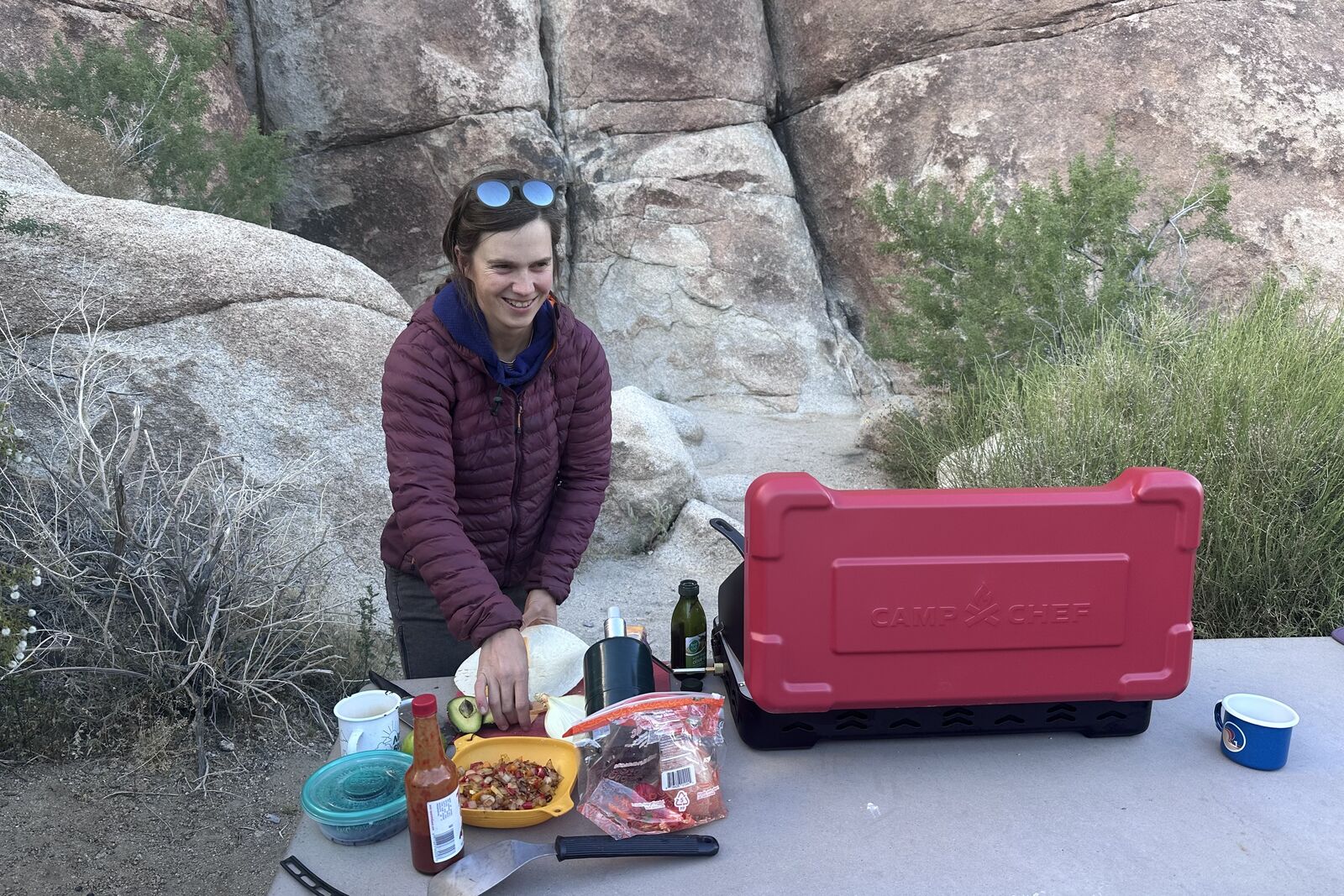 A woman smiles while preparing quesadillas on a picnic table next to a large double-burner stove in the desert.