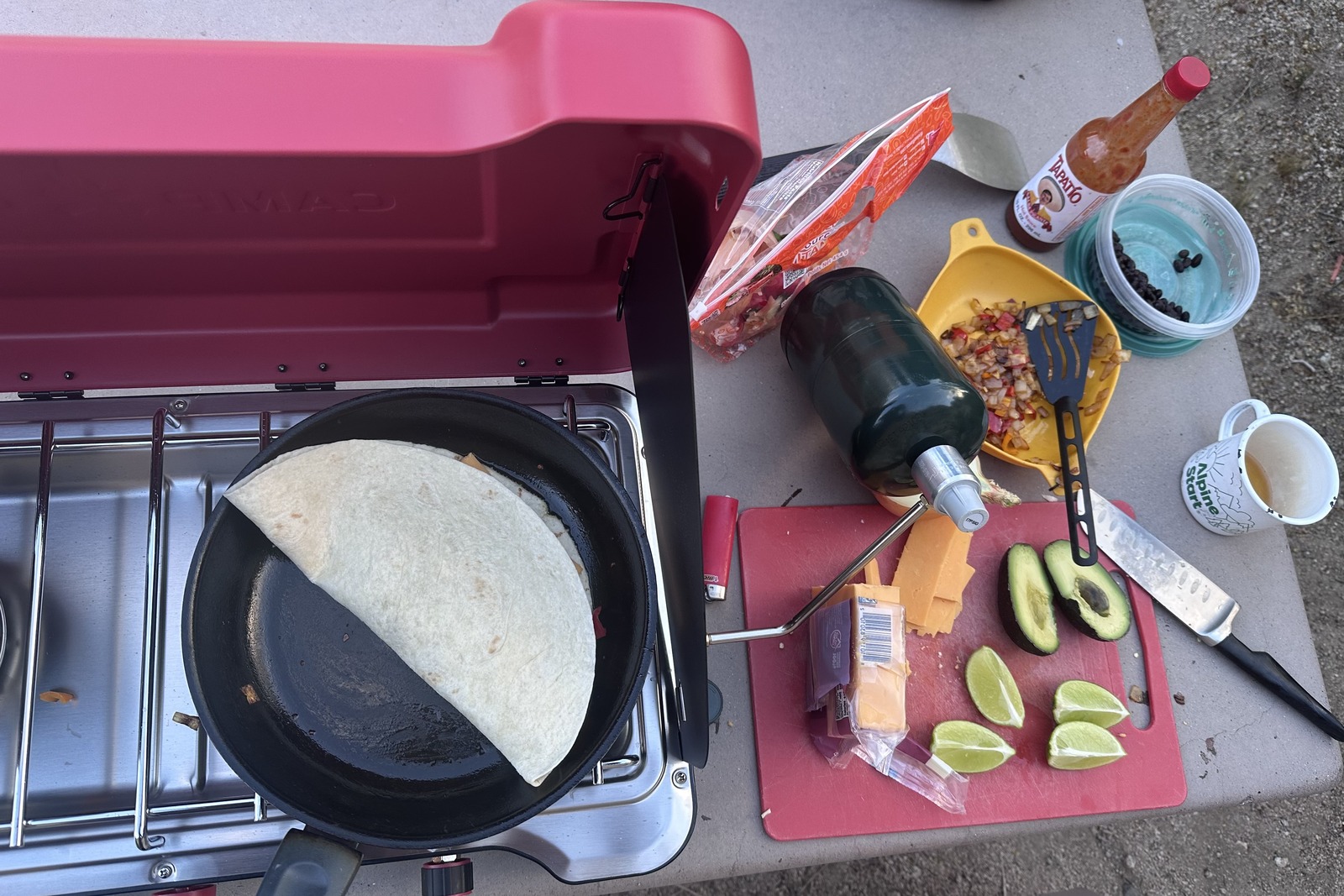 Top down / close-up of one side of a double-burner stove holding a frying pan with a quesadilla. A cutting board next to the stove has sliced cheese, avocado, and limes.