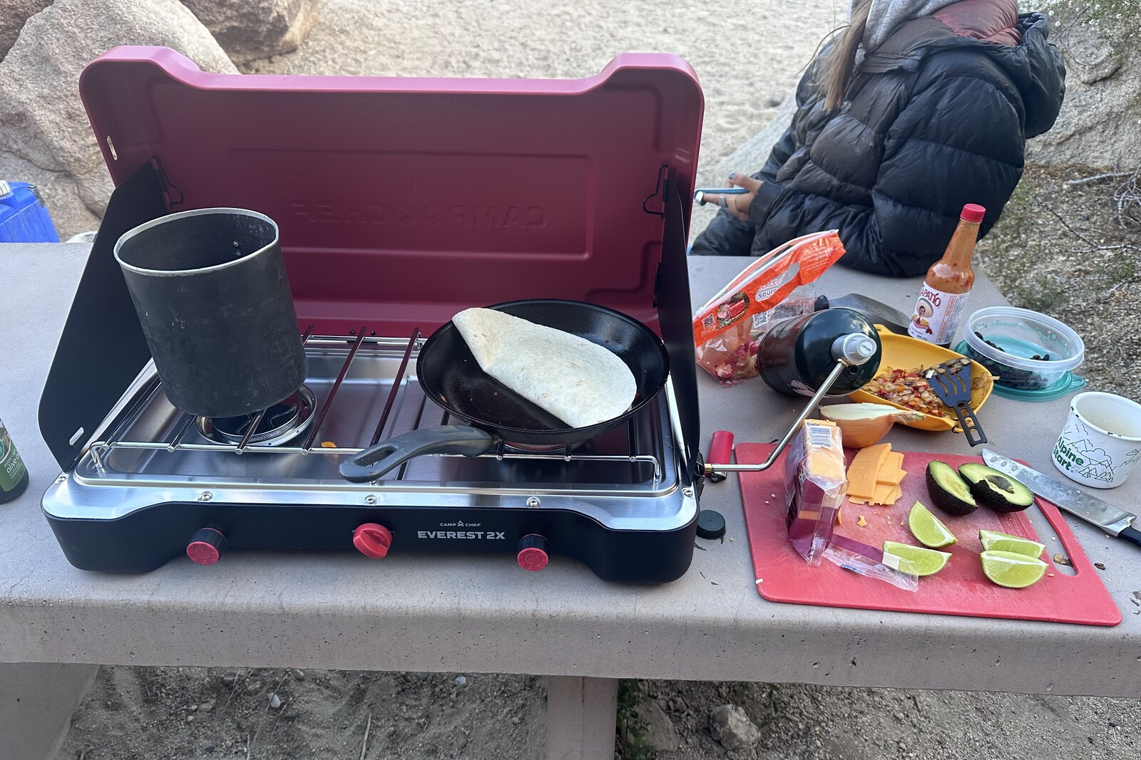 Close-up of a picnic table with a red double burner stove on top. A cutting board with cheese, avacado, and limes sits next to the stove, and a quesadilla cooks on a pan on top of the stove.