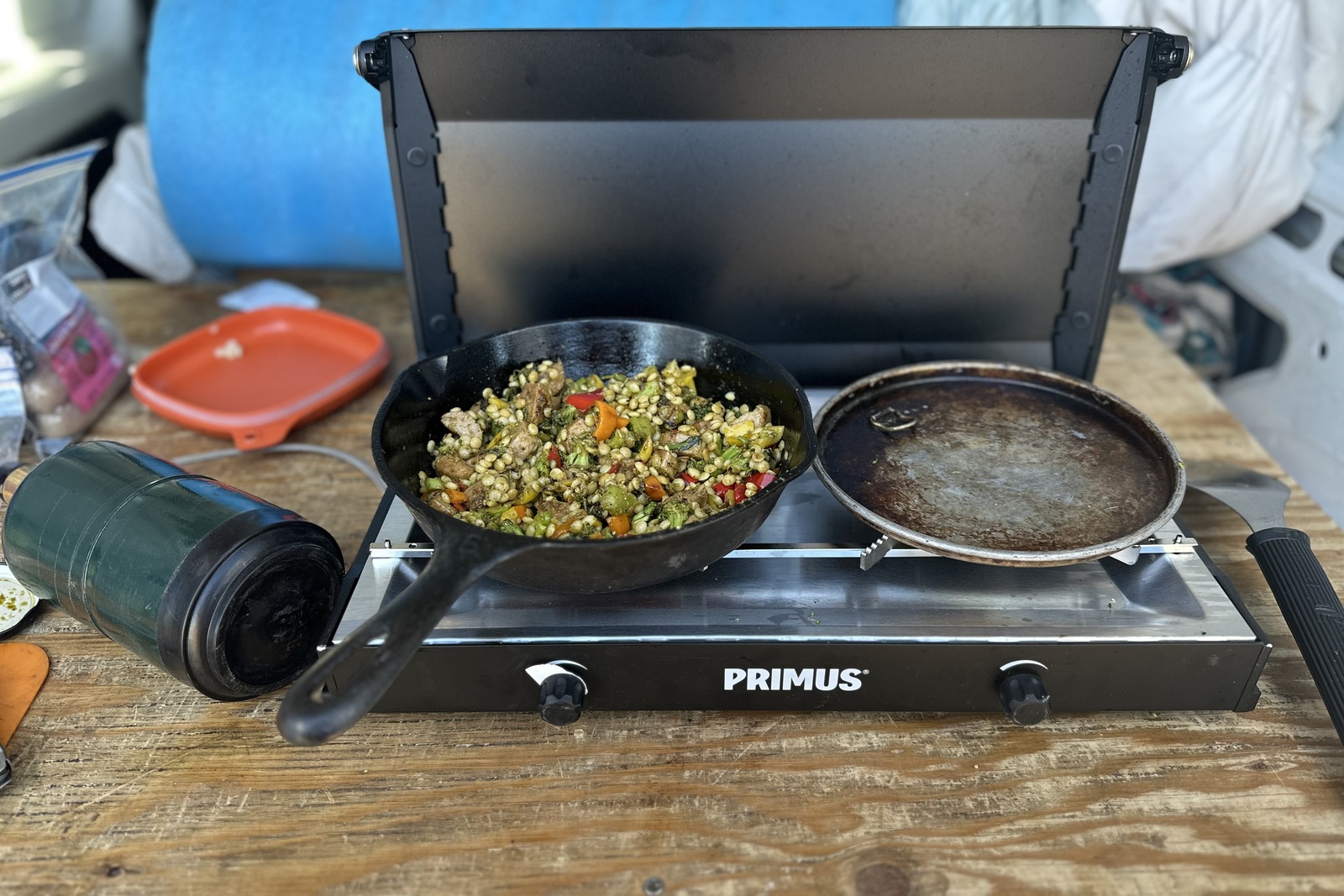 Close-up image of a small, black double-burner stove. On one of the burners, a small cast iron skillet holds veggies, chicken sausage, and couscous.