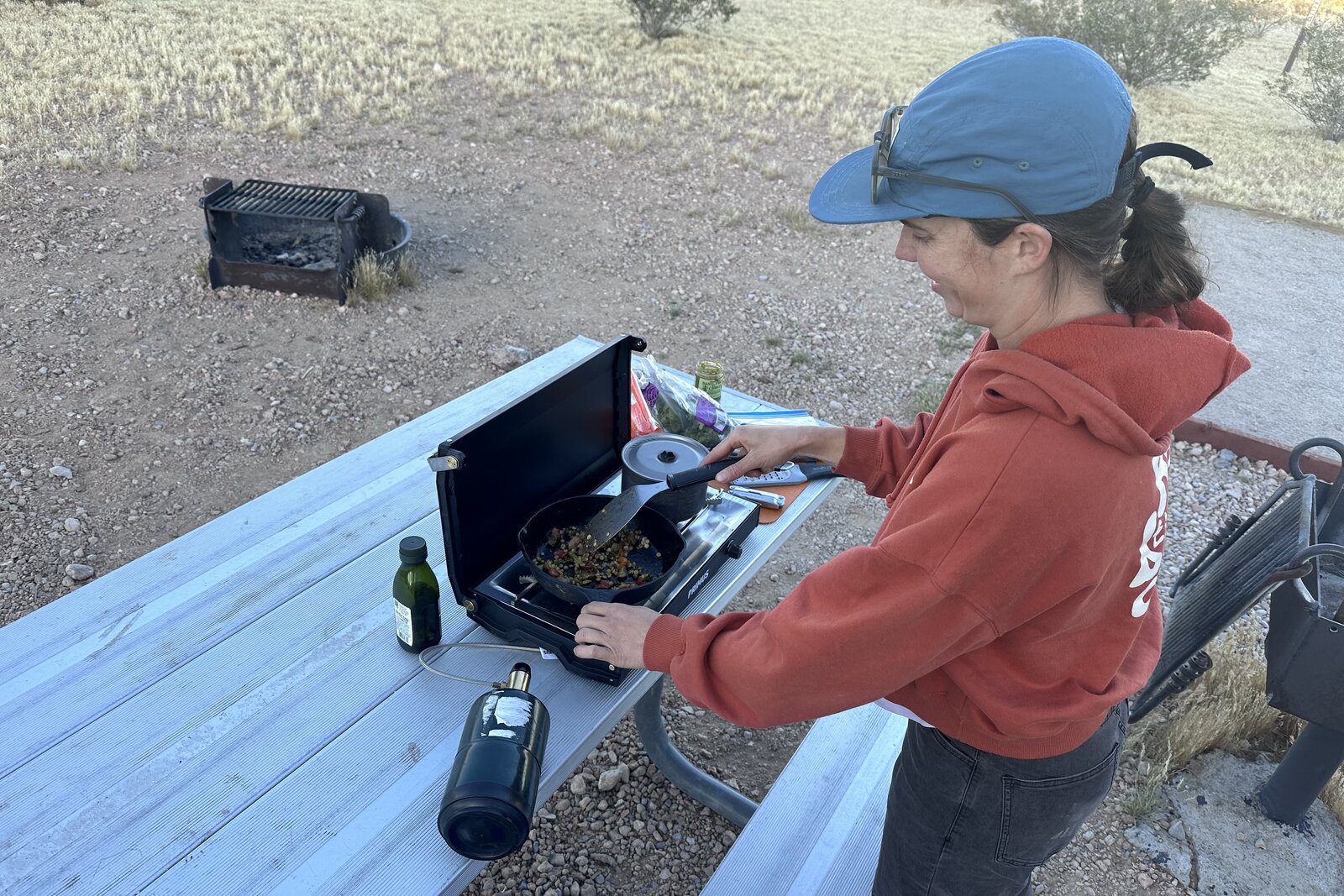 A woman in a red sweatshirt stirs food in a pan on a small, black double-burner stove.