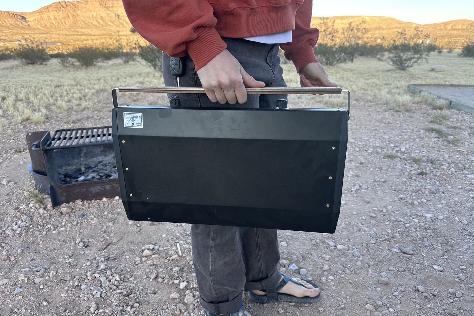 Close-up image of a woman carrying a slim, double-burner stove with an attached wooden handle.