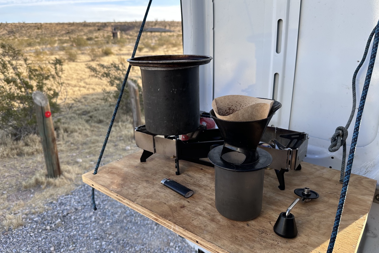 Close-up image of a small double-burner stove with a pot of water on one of the burners. The stove rests on top of a wooden fold-out table attached to the back door of a white van.