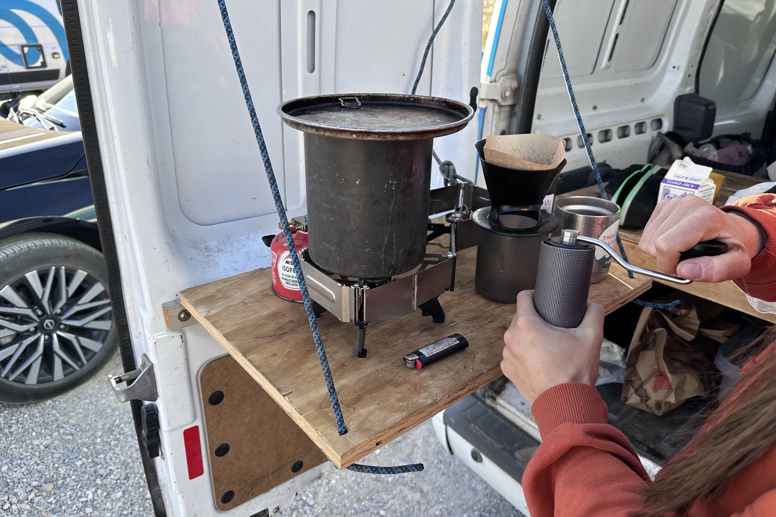 Close-up of a woman grinding coffee with a hand grinder in front of a small double-burner stove sitting on top of a wooden fold-out table on the back door of a van.