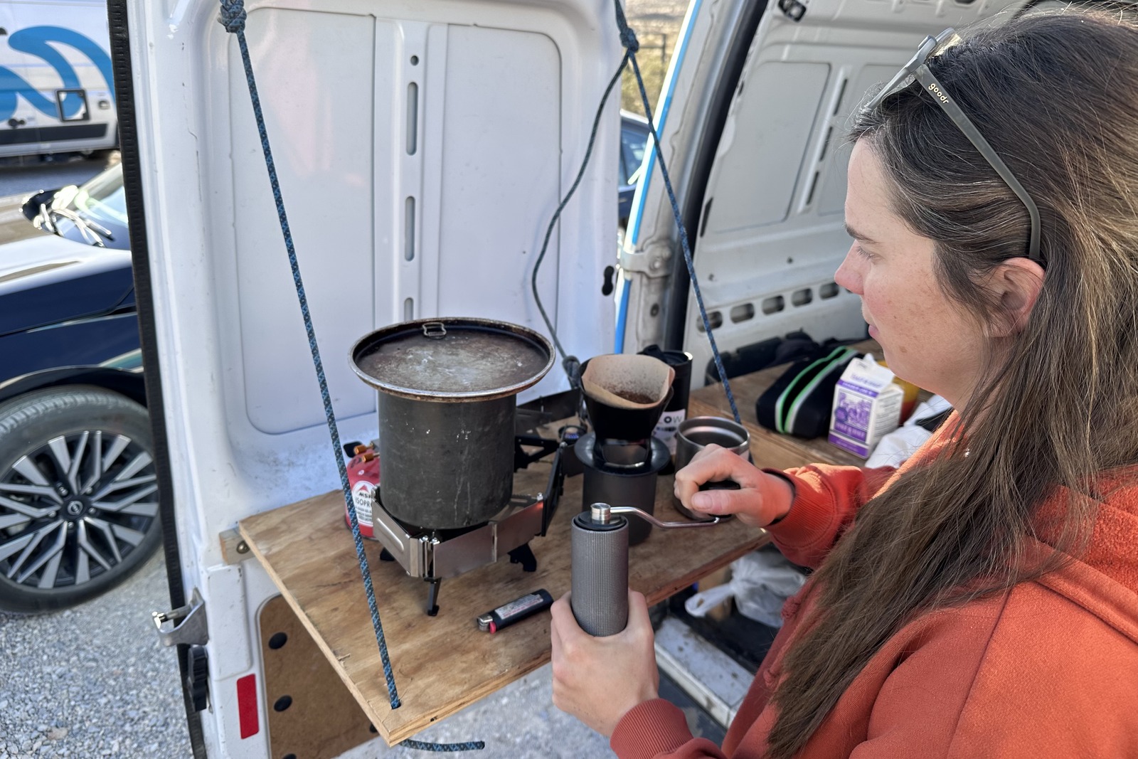 A woman grinds coffee with a hand grinder while a pot of water boils on a small double-burner stove sitting on a wooden fold-out table attached to the back door of a white van.
