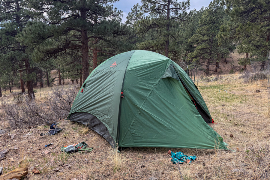 The Kelty Wireless 6 tent pitched at a forest campsite with the rainfly fully deployed and the front vestibule closed.