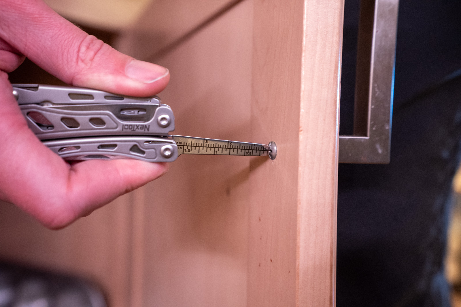 A hand holds a multi tool with a screwdriver extended fitting into a screw on a cabinet