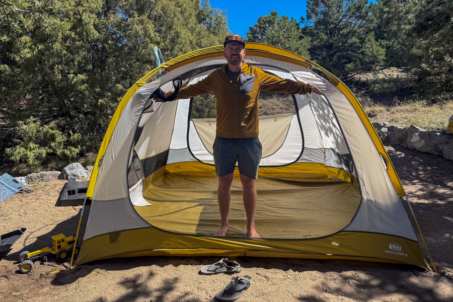 A man stands inside the front opening of the REI Base Camp 6 tent to show the tent’s interior width and standing height.
