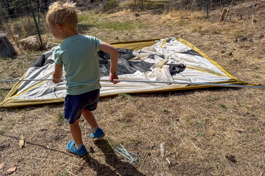 A child helps set up the REI Base Camp 6 tent by carrying a long aluminum tent pole across camp.