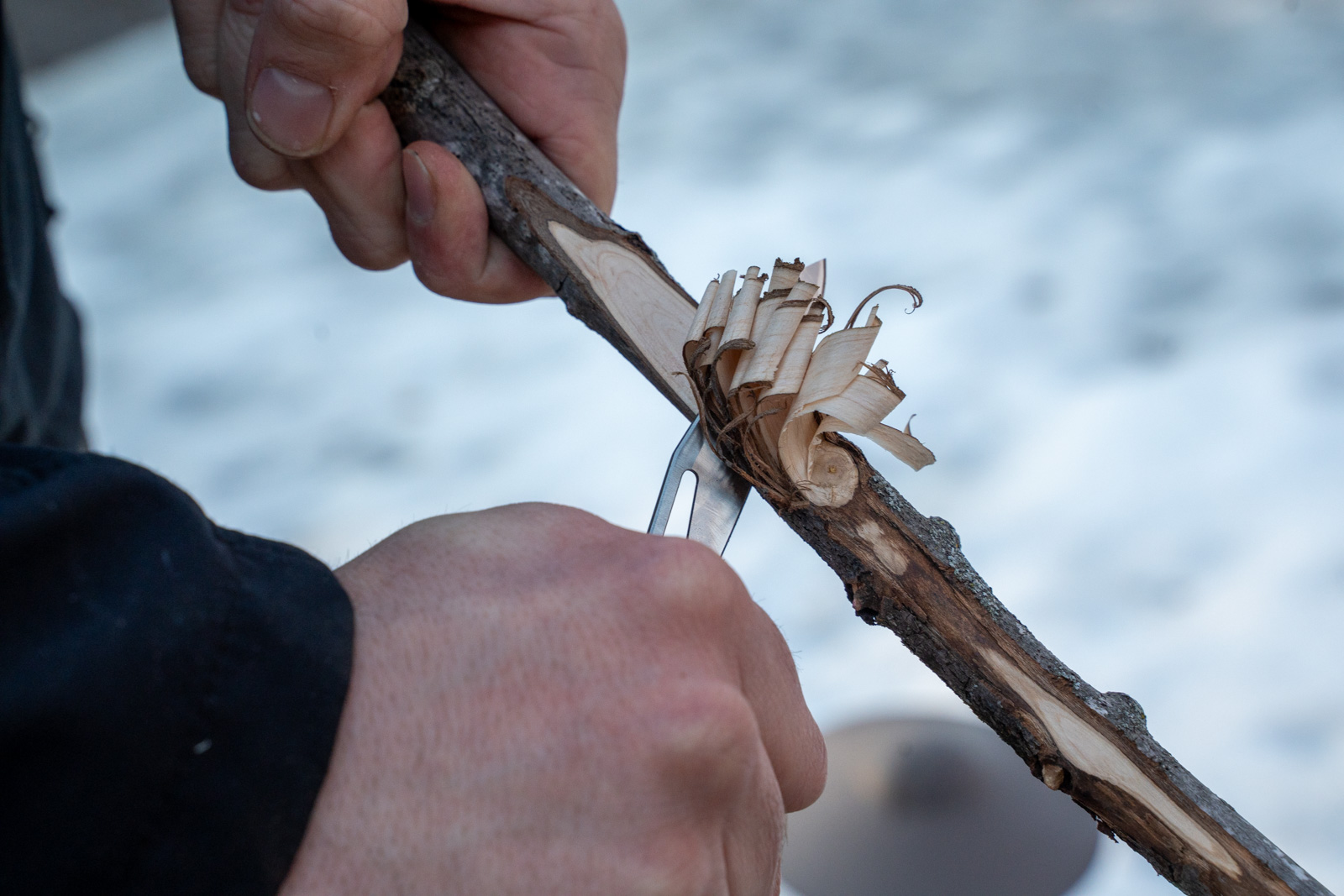 A man shaves curls of wood from a stick with a knife