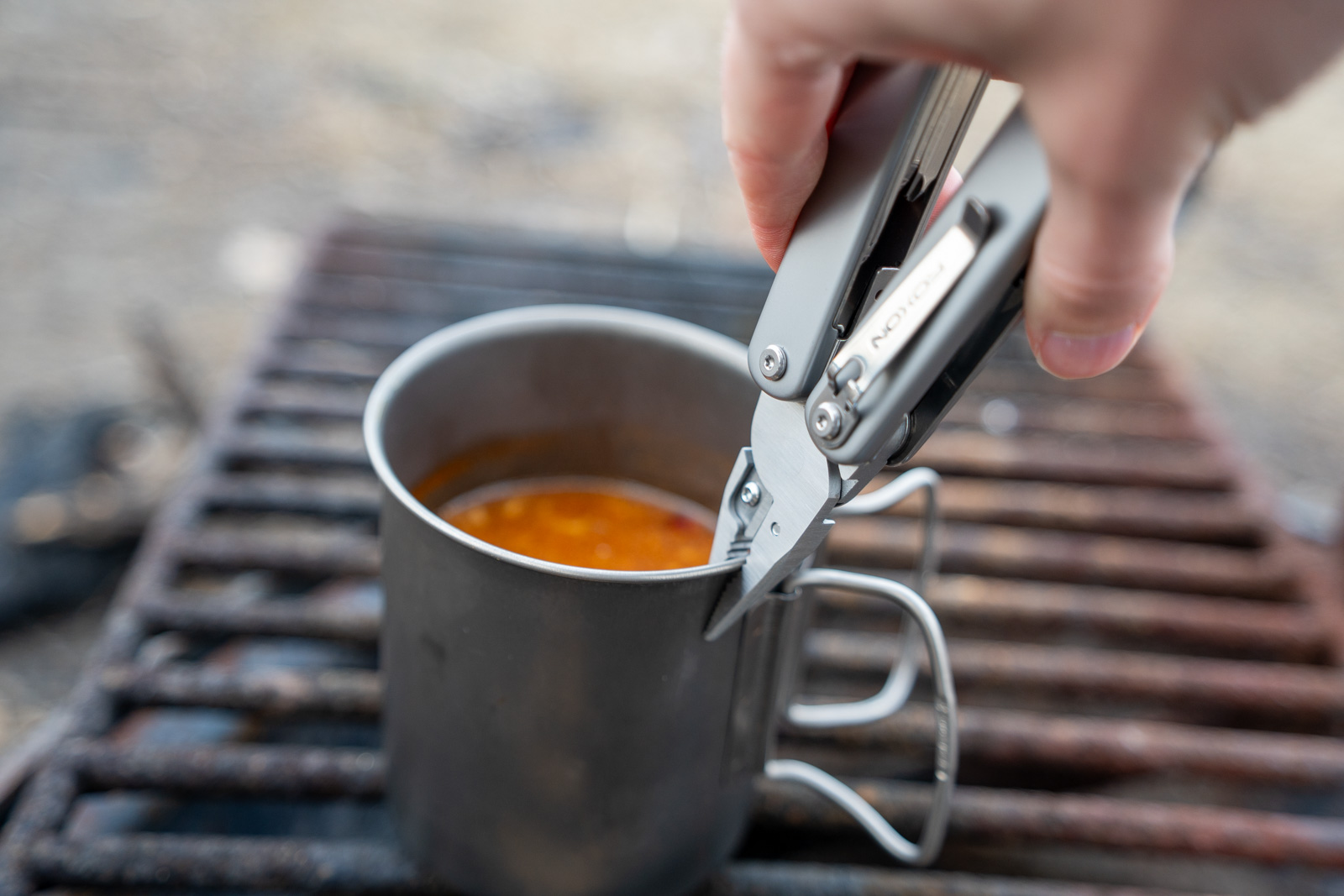 A hand holds a multi tool gripping a cup with the pliers over a campfire grate