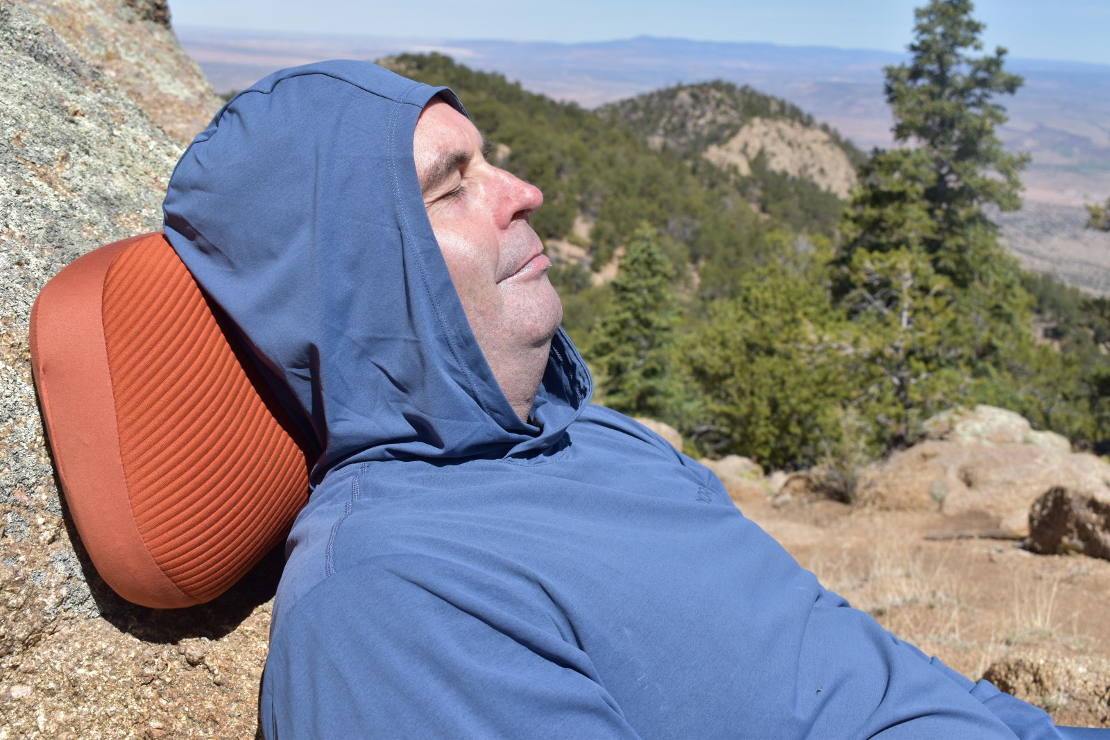 A hiker reclines against a cliff with an orange Aeros Premium pillow cushioning his head.