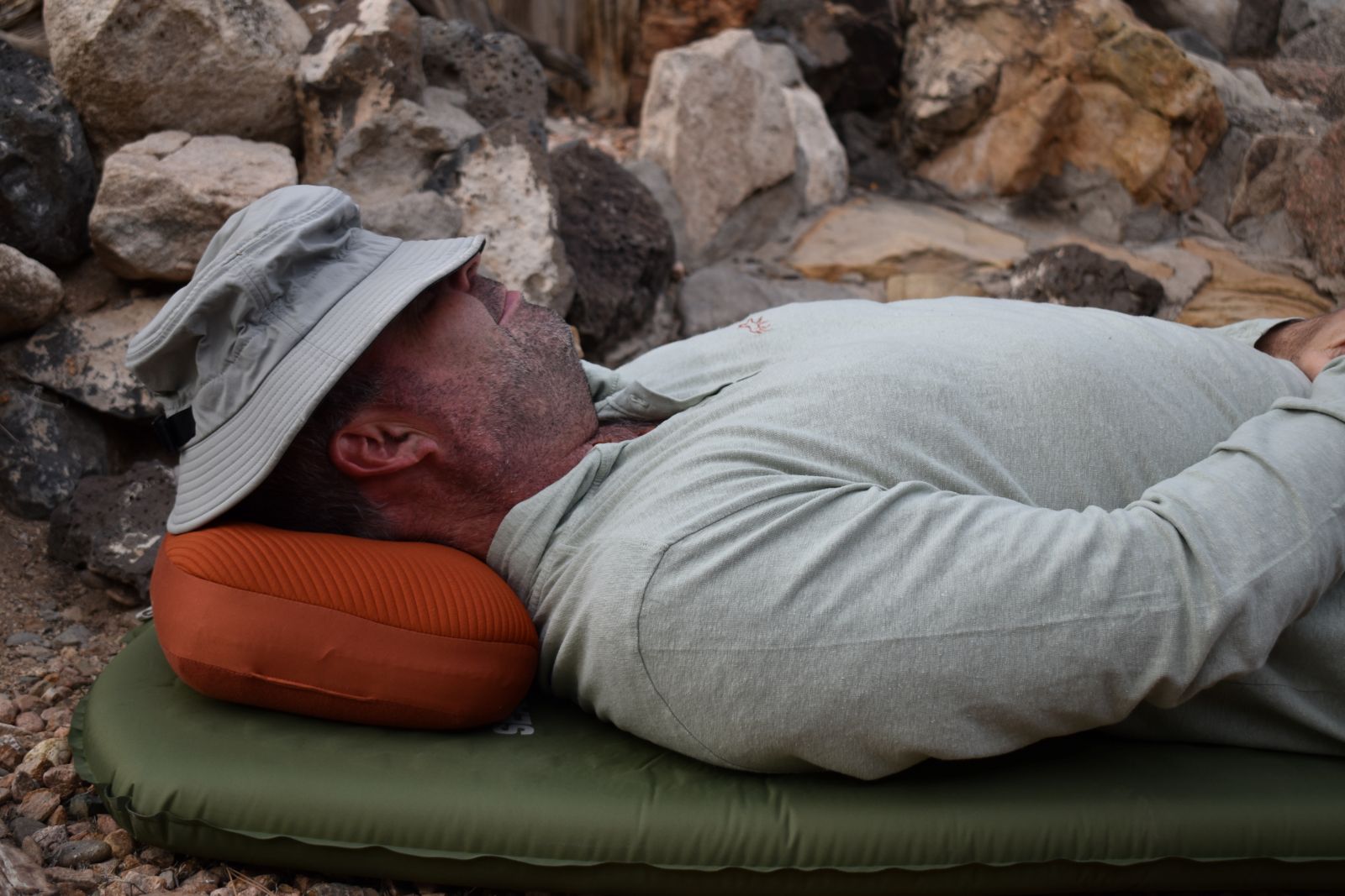 A hiker lies down on the Aeros Premium Pillow with a bucket hat pulled over his eyes.