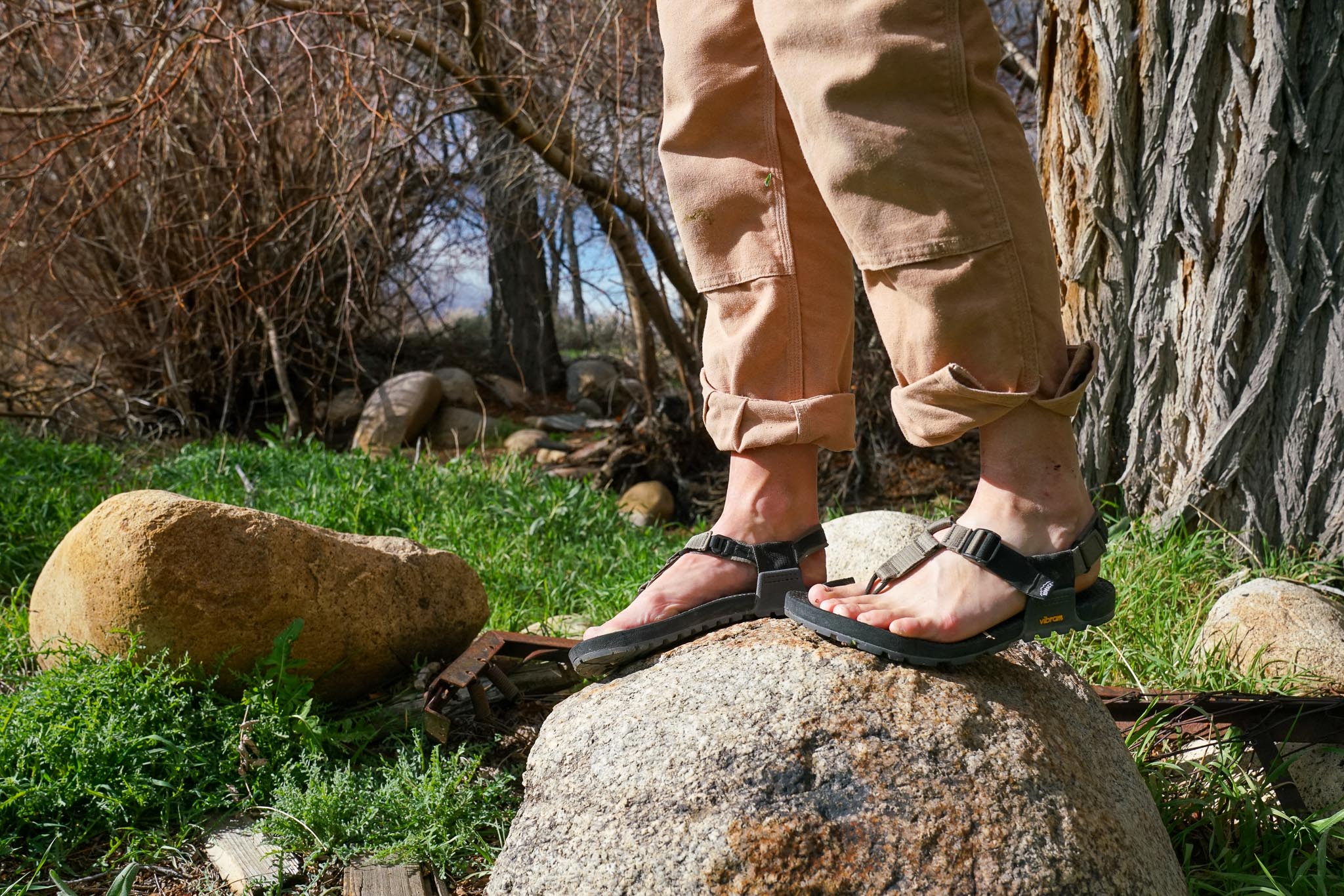 A close up of a person's feet wearing sandals standing on a boulder with trees and grass in the background.