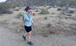 A hiker walks on a sandy trail in the desert while drinking out of a hydration hose.