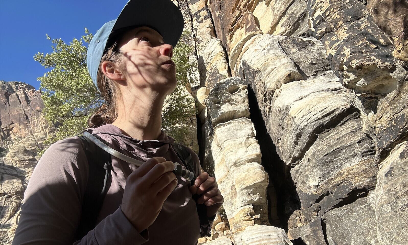 A hiker looks up at a cliff while holding a hydration hose