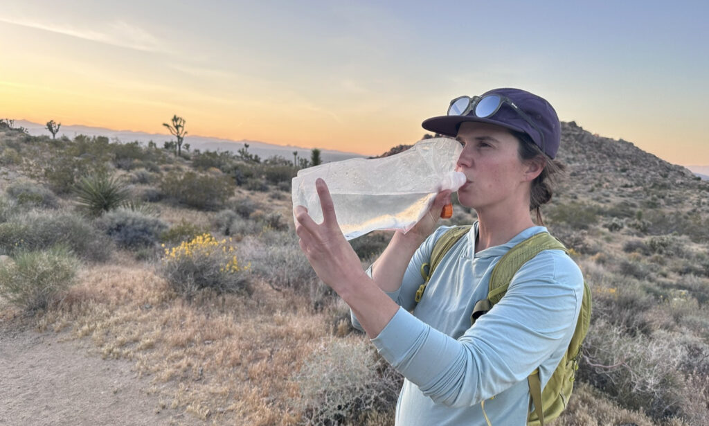 A hiker drinks from a water bladder without a hose. A desert sunset is in the background.