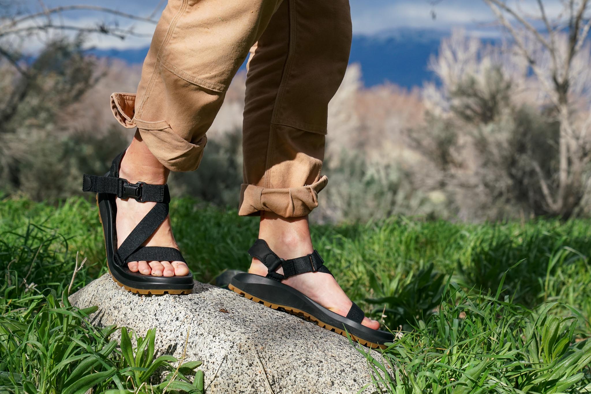 A close up of a person's feet wearing sandals standing on a rock amidst grass.