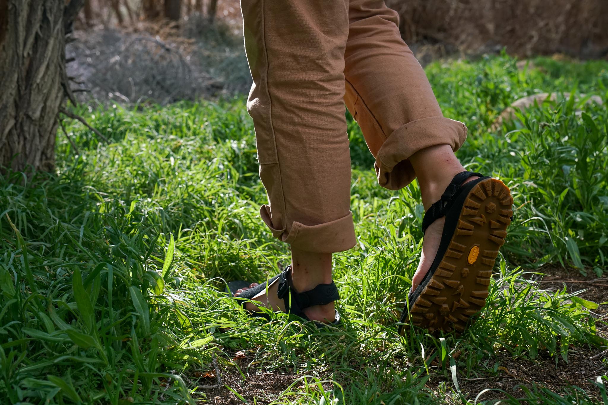 A close up of a person's feet wearing sandals and walking on grass. They walk away from the camera, showing the underside of one sandal.