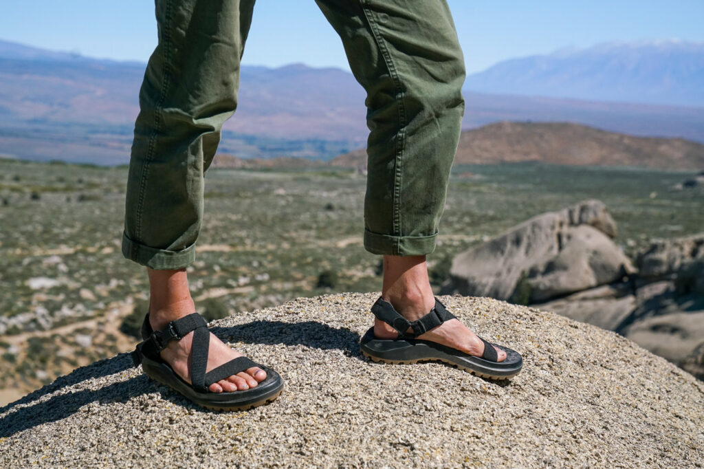 A close up of a person's feet wearing sandals standing on a rock. There are mountains in the background.