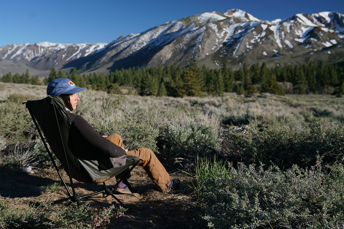 A person sits in a scoop-seat camping chair with mountains in the background.