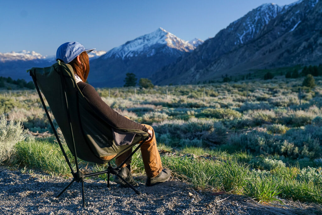 A person sits with their back to the camera in a scoop-seat camping chair with mountains in the background.