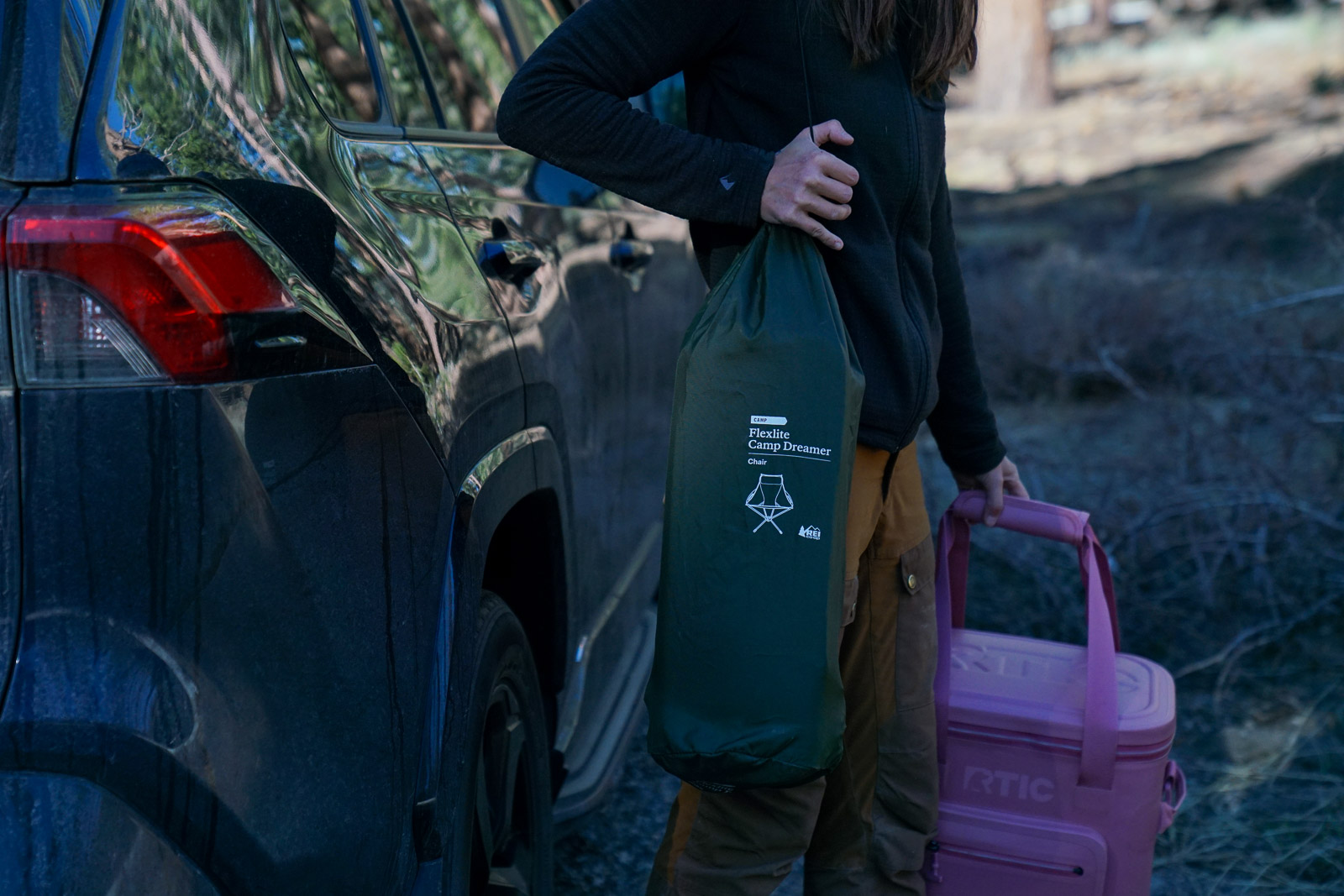 A close up of a person walking away from a car with a cooler in one hand and a camping chair stowed in a stuff sack over one shoulder.